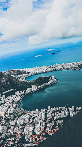 Aerial view of a coastal city with a large lagoon bordered by urban development. High-rise buildings and residential areas are nestled between lush green mountains and the ocean. The sky is partly cloudy, with patches of blue peeking through.