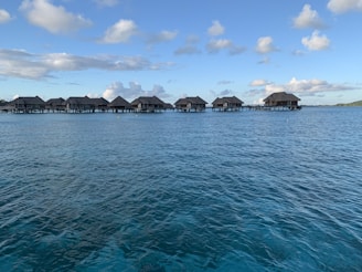 A series of overwater bungalows extend across a calm, clear blue sea under a sky dotted with fluffy clouds. The bungalows have thatched roofs and are connected by a wooden walkway.