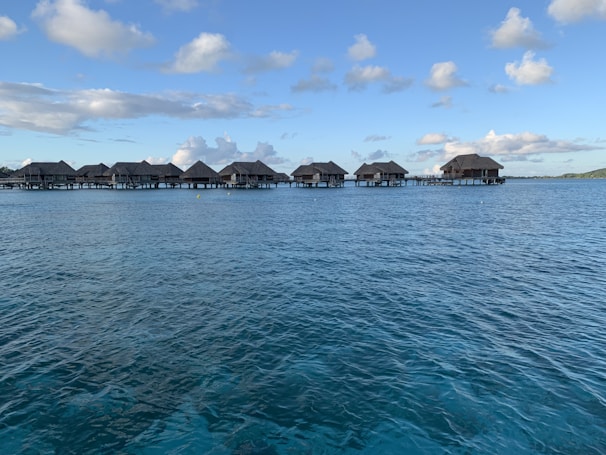 A series of overwater bungalows extend across a calm, clear blue sea under a sky dotted with fluffy clouds. The bungalows have thatched roofs and are connected by a wooden walkway.