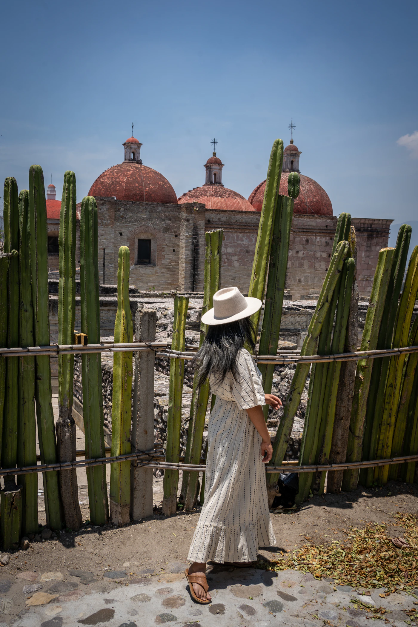 A woman in a white wedding dress photographed at Mitla archaeological ruins in Oaxaca Mexico with cactus fence and red domes visible behind her