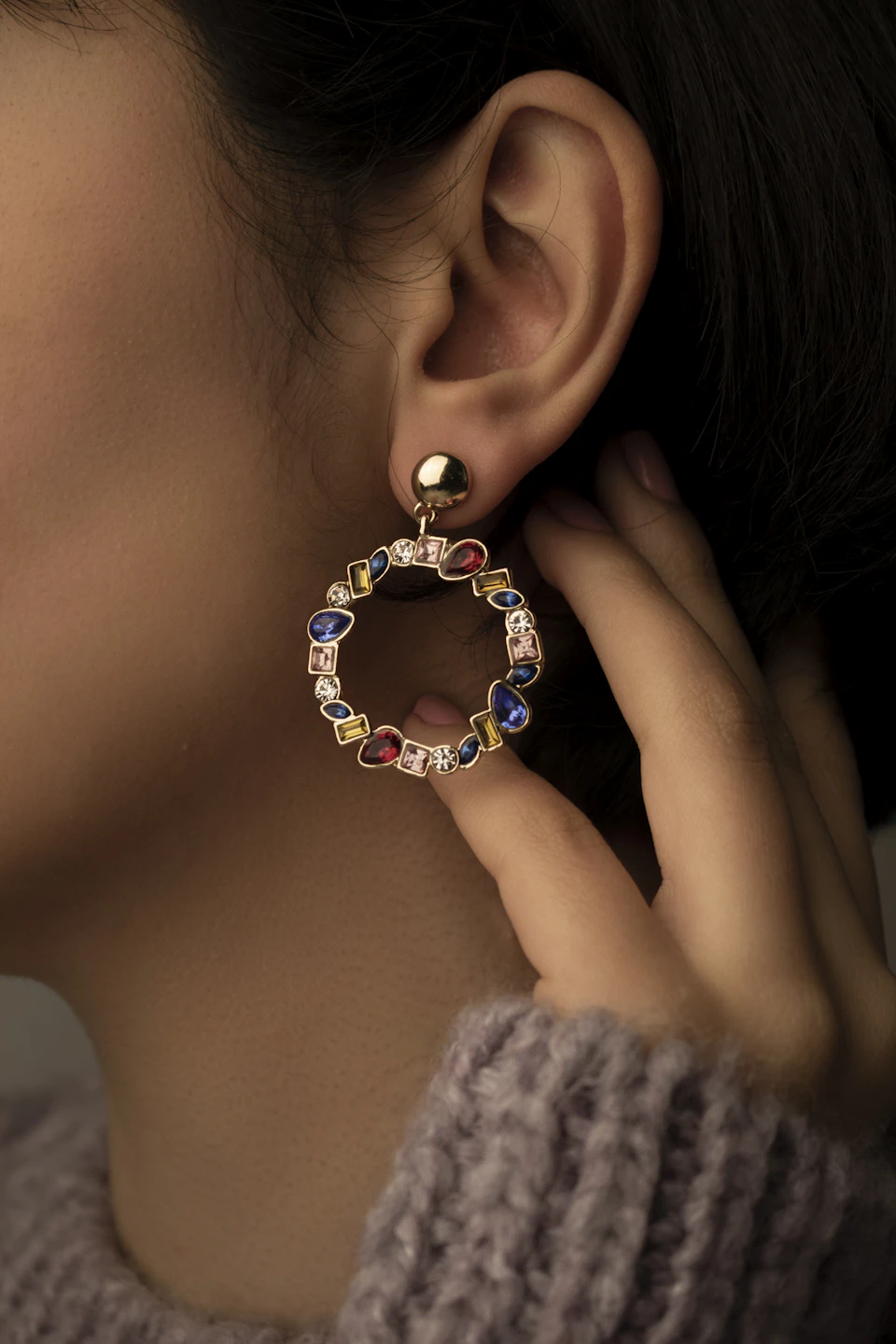 The image shows a close-up view of a person's ear, with their left ear adorned with a pair of colorful diamond earrings. The earrings are encircled by a gold ring and feature a vibrant mix of blue, green, yellow, and red stones. The person wearing the earrings is positioned in front of a gray background, which contrasts with the bright colors of the earrings and draws attention to them.