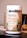 A container labeled 'Youthful Living Keto Vegan Protein Shake' is prominently displayed. The container highlights its plant-based ingredients and is flavor 'Cafe Mocca'. It's positioned on a reflective surface with part of a banana visible in the background.