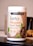A container labeled 'Youthful Living Keto Vegan Protein Shake' is prominently displayed. The container highlights its plant-based ingredients and is flavor 'Cafe Mocca'. It's positioned on a reflective surface with part of a banana visible in the background.