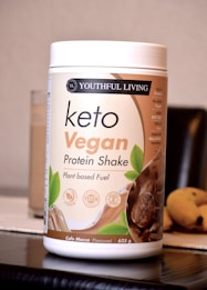 A container labeled 'Youthful Living Keto Vegan Protein Shake' is prominently displayed. The container highlights its plant-based ingredients and is flavor 'Cafe Mocca'. It's positioned on a reflective surface with part of a banana visible in the background.