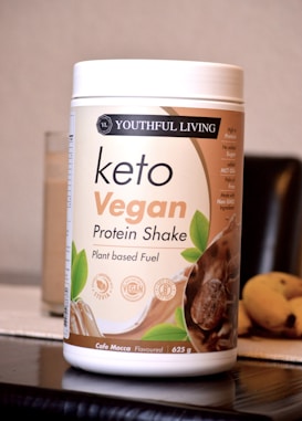 A container labeled 'Youthful Living Keto Vegan Protein Shake' is prominently displayed. The container highlights its plant-based ingredients and is flavor 'Cafe Mocca'. It's positioned on a reflective surface with part of a banana visible in the background.