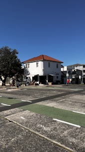 A two-story building with a red-tiled roof stands on a quiet street. The building is painted white and has several windows. It is surrounded by a clear blue sky and a tree on the left side, casting shadows on the facade. There is a small outdoor seating area and a sign on the sidewalk indicating a business establishment.