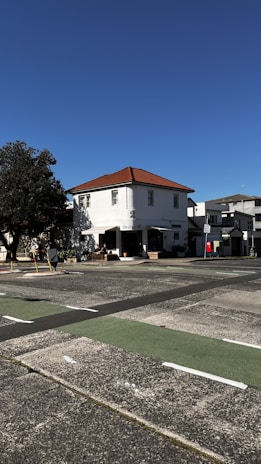 A two-story building with a red-tiled roof stands on a quiet street. The building is painted white and has several windows. It is surrounded by a clear blue sky and a tree on the left side, casting shadows on the facade. There is a small outdoor seating area and a sign on the sidewalk indicating a business establishment.