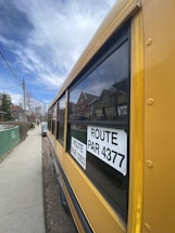 A yellow school bus is parked on a residential street. The bus has large windows with black frames and two signs displaying the route numbers 3303 and 4377. Houses with pitched roofs are visible in the background, and a couple of cars are parked along the sidewalk.