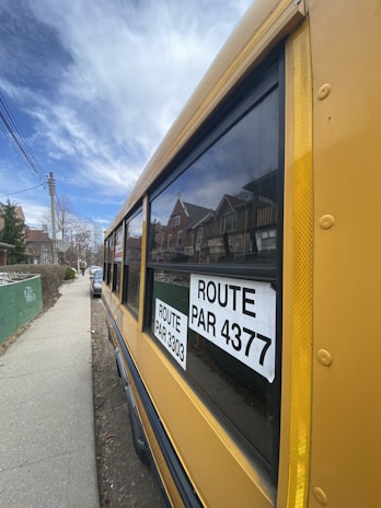 A yellow school bus is parked on a residential street. The bus has large windows with black frames and two signs displaying the route numbers 3303 and 4377. Houses with pitched roofs are visible in the background, and a couple of cars are parked along the sidewalk.
