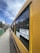A yellow school bus is parked on a residential street. The bus has large windows with black frames and two signs displaying the route numbers 3303 and 4377. Houses with pitched roofs are visible in the background, and a couple of cars are parked along the sidewalk.
