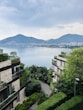 Luxurious residential buildings surrounded by lush greenery with a view of a tranquil bay and distant cityscape under a cloudy sky. Mountains are visible in the background, enhancing the scenic beauty of the area.