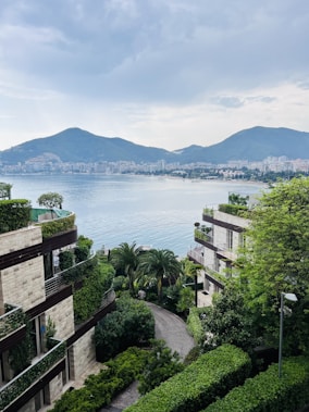 Luxurious residential buildings surrounded by lush greenery with a view of a tranquil bay and distant cityscape under a cloudy sky. Mountains are visible in the background, enhancing the scenic beauty of the area.