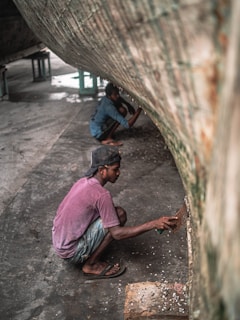 Two individuals are working underneath the hull of a large boat, scraping away at the surface. One is closer to the camera, wearing a cap, a purple shirt, and jeans shorts, using a scraping tool. The other person is in the background, dressed in blue, focused on a similar task. The environment appears industrial, with a concrete floor, and the underside of the boat dominates the frame.