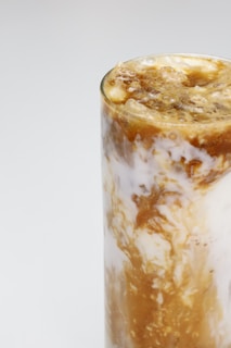 A tall glass filled with a swirled mixture of creamy white and rich caramel brown liquid, possibly a coffee beverage with milk or cream. The texture appears frothy with visible layers.