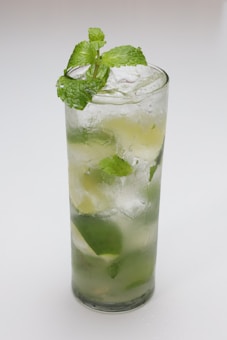A tall glass filled with a refreshing cocktail, featuring ice cubes, lime wedges, and fresh mint leaves. The glass is frosted and contains a transparent, fizzy liquid. A sprig of mint decorates the top of the drink.