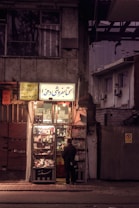 A small, dimly lit bookstore exterior is depicted. The shop is filled with various items such as books and magazines displayed at the entrance. A person stands in front, browsing the collection. The signs above the entrance are written in Persian script. The scene is set during twilight, creating a moody and atmospheric vibe.