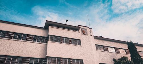 A building with a textured white facade and multiple rectangular windows featuring brown accents. The structure has a flat roof and an emblem or shield near the top. A clear blue sky with scattered clouds serves as the backdrop. A light pole is visible on the roof, and some greenery is present in the foreground.