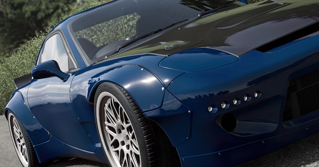 A sleek, modified sports car with a shiny blue exterior and white rims is parked or moving slowly on an asphalt road. The car features a carbon fiber hood and a distinct aerodynamic design, complemented by lush green foliage in the background that contrasts with the car's polished surface.