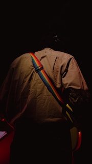 A person stands with their back to the camera, wearing a beige jacket and a colorful rainbow guitar strap. The background is dark and the person appears to be playing a guitar, which is partially visible.