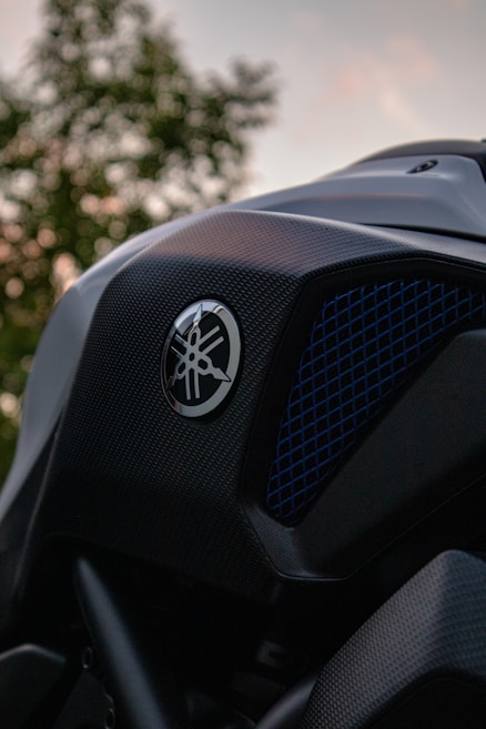 Close-up view of a motorcycle's body with a focus on the Yamaha logo and textured black surface. The background features an out-of-focus tree against the sky during sunset.