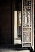 A detailed wooden door with intricate carvings and lattice design is partially open. The door is part of a larger structure with dark wooden walls and floors. Light from the outside illuminates part of the interior, casting soft shadows and highlighting the craftsmanship of the woodwork.