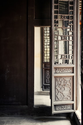 A detailed wooden door with intricate carvings and lattice design is partially open. The door is part of a larger structure with dark wooden walls and floors. Light from the outside illuminates part of the interior, casting soft shadows and highlighting the craftsmanship of the woodwork.
