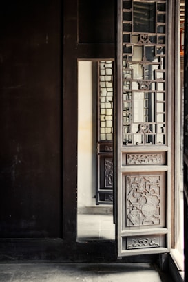 A detailed wooden door with intricate carvings and lattice design is partially open. The door is part of a larger structure with dark wooden walls and floors. Light from the outside illuminates part of the interior, casting soft shadows and highlighting the craftsmanship of the woodwork.