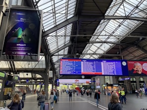 A busy train station interior with a large digital display board showing train schedules. Several travelers are walking or standing with luggage. The station's architecture includes a high, industrial-style metal ceiling with large glass panels allowing natural light to enter. An advertisement for a mobile phone is visible on a screen to the left, while a digital clock and other advertisements are positioned near the schedule board.