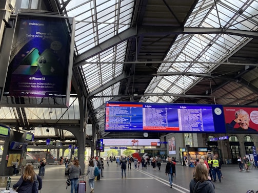 A busy train station interior with a large digital display board showing train schedules. Several travelers are walking or standing with luggage. The station's architecture includes a high, industrial-style metal ceiling with large glass panels allowing natural light to enter. An advertisement for a mobile phone is visible on a screen to the left, while a digital clock and other advertisements are positioned near the schedule board.