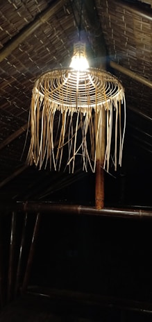 A bamboo woven lampshade hangs from a thatched roof, with warm light illuminating the structure. The roof and lamp are constructed from natural materials, emitting a rustic and traditional vibe.
