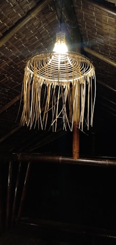 A bamboo woven lampshade hangs from a thatched roof, with warm light illuminating the structure. The roof and lamp are constructed from natural materials, emitting a rustic and traditional vibe.