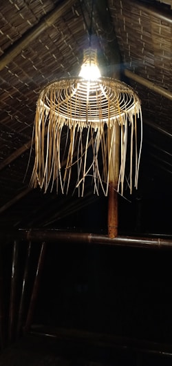 A bamboo woven lampshade hangs from a thatched roof, with warm light illuminating the structure. The roof and lamp are constructed from natural materials, emitting a rustic and traditional vibe.
