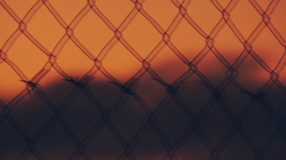 A chain-link fence with barbed wire is silhouetted against a vibrant orange sky, creating a striking contrast. The background is blurred, enhancing the focus on the fence pattern.
