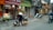 A street scene featuring a person wearing a conical hat, carrying goods in a traditional yoke. The background includes shops with colorful signs and displays of fruits and vegetables. Motorbikes are visible on the street alongside the pedestrian.