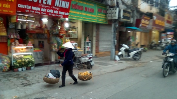 A street scene featuring a person wearing a conical hat, carrying goods in a traditional yoke. The background includes shops with colorful signs and displays of fruits and vegetables. Motorbikes are visible on the street alongside the pedestrian.