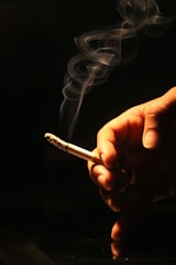 A hand holding a lit cigarette with smoke elegantly swirling upwards against a dark background, highlighting the cigarette's glowing tip and the warm tones of the skin.