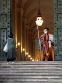 A person in a colorful striped uniform stands guard at the entrance of a grand, arched building. Another person, carrying a bag, faces the guard and seems to be approaching them. The setting is elegant and historic, with large doors and illuminated lamps along the walls.