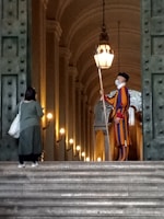 A person in a colorful striped uniform stands guard at the entrance of a grand, arched building. Another person, carrying a bag, faces the guard and seems to be approaching them. The setting is elegant and historic, with large doors and illuminated lamps along the walls.