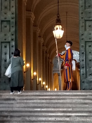 A person in a colorful striped uniform stands guard at the entrance of a grand, arched building. Another person, carrying a bag, faces the guard and seems to be approaching them. The setting is elegant and historic, with large doors and illuminated lamps along the walls.