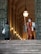 A person in a colorful striped uniform stands guard at the entrance of a grand, arched building. Another person, carrying a bag, faces the guard and seems to be approaching them. The setting is elegant and historic, with large doors and illuminated lamps along the walls.