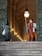 A person in a colorful striped uniform stands guard at the entrance of a grand, arched building. Another person, carrying a bag, faces the guard and seems to be approaching them. The setting is elegant and historic, with large doors and illuminated lamps along the walls.