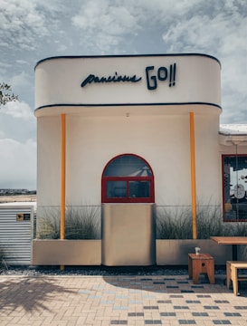 A modern building exterior with a minimalist design featuring a smooth, rounded structure. A bold sign reads 'Go!!' above a red arched window flanked by bright orange columns. The facade is adorned with tall ornamental grasses. A wooden table and a small stool sit on a patterned brick patio in front of the building.