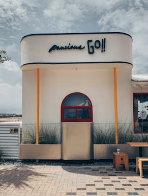 A modern building exterior with a minimalist design featuring a smooth, rounded structure. A bold sign reads 'Go!!' above a red arched window flanked by bright orange columns. The facade is adorned with tall ornamental grasses. A wooden table and a small stool sit on a patterned brick patio in front of the building.