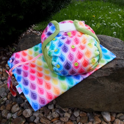 A colorful reusable cloth diaper and matching wet bag featuring a vibrant rainbow leaf pattern with green borders are placed on a rock outside on a bed of small stones. The background shows a green grassy area with some white flowers.