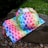 A colorful reusable cloth diaper and matching wet bag featuring a vibrant rainbow leaf pattern with green borders are placed on a rock outside on a bed of small stones. The background shows a green grassy area with some white flowers.