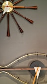 A modern, artistic clock featuring a central gold face with elongated brown spokes extending outward. Below is a curved, wave-like element with a dark circular base and geometric clock hands in white and orange.