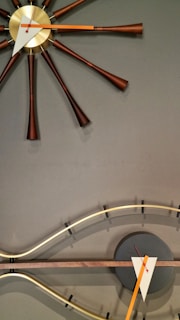 A modern, artistic clock featuring a central gold face with elongated brown spokes extending outward. Below is a curved, wave-like element with a dark circular base and geometric clock hands in white and orange.