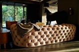 A luxurious room features a large, tufted, beige sofa with a gray blanket draped over it. A dark horse-shaped lamp stands next to an end table, creating a striking decorative element. Floor-to-ceiling windows reveal greenery outside, allowing natural light to illuminate the space. The interior is stylishly furnished with modern elements and a rich color palette.