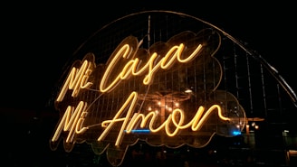 A neon sign displaying the words 'Mi Casa Mi Amor' in a warm yellow glow against a dark background. The sign is mounted in front of a metal grid, adding an industrial feel to the romantic message.