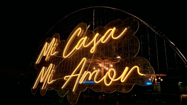 A neon sign displaying the words 'Mi Casa Mi Amor' in a warm yellow glow against a dark background. The sign is mounted in front of a metal grid, adding an industrial feel to the romantic message.